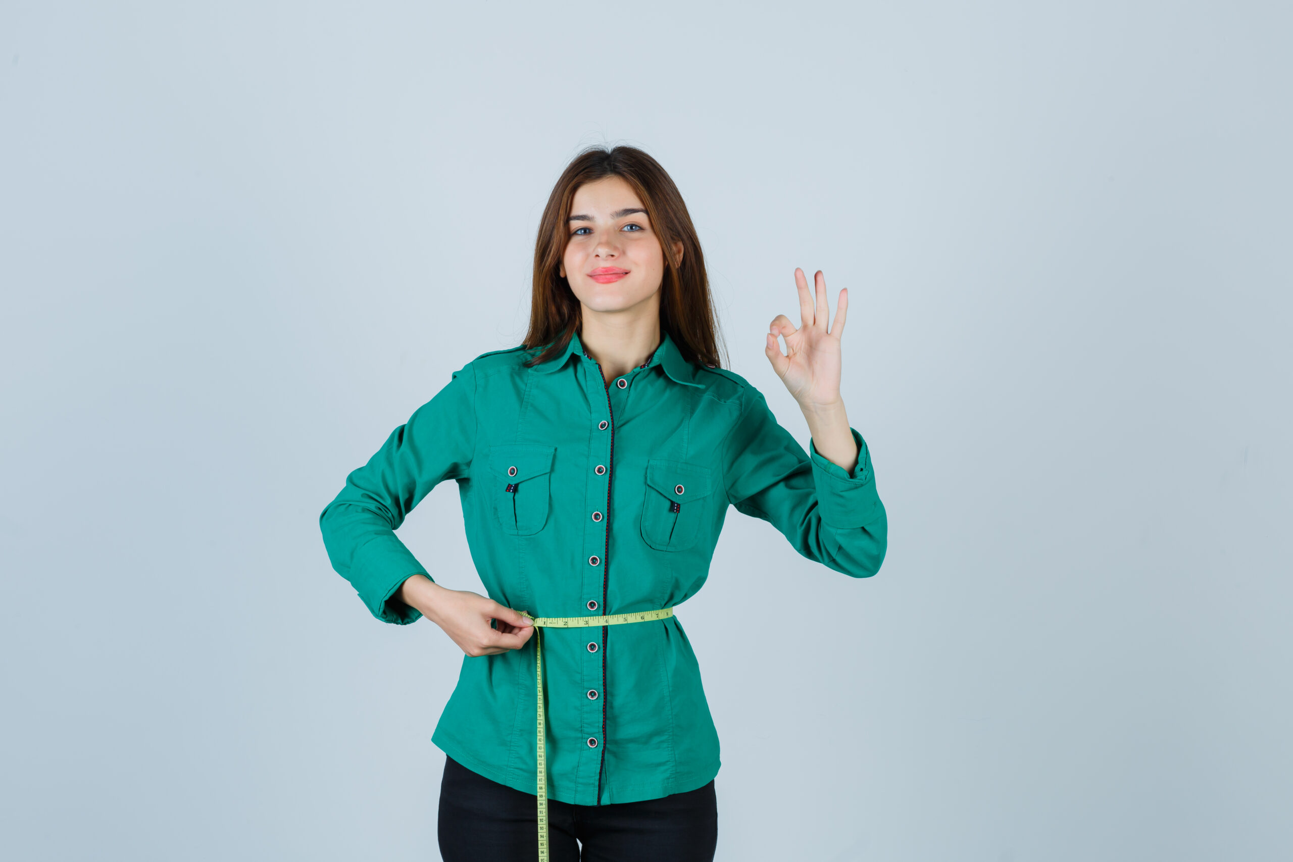 young lady holding measuring tape around her waist while showing ok gesture in shirt, pants and looking glad. front view.